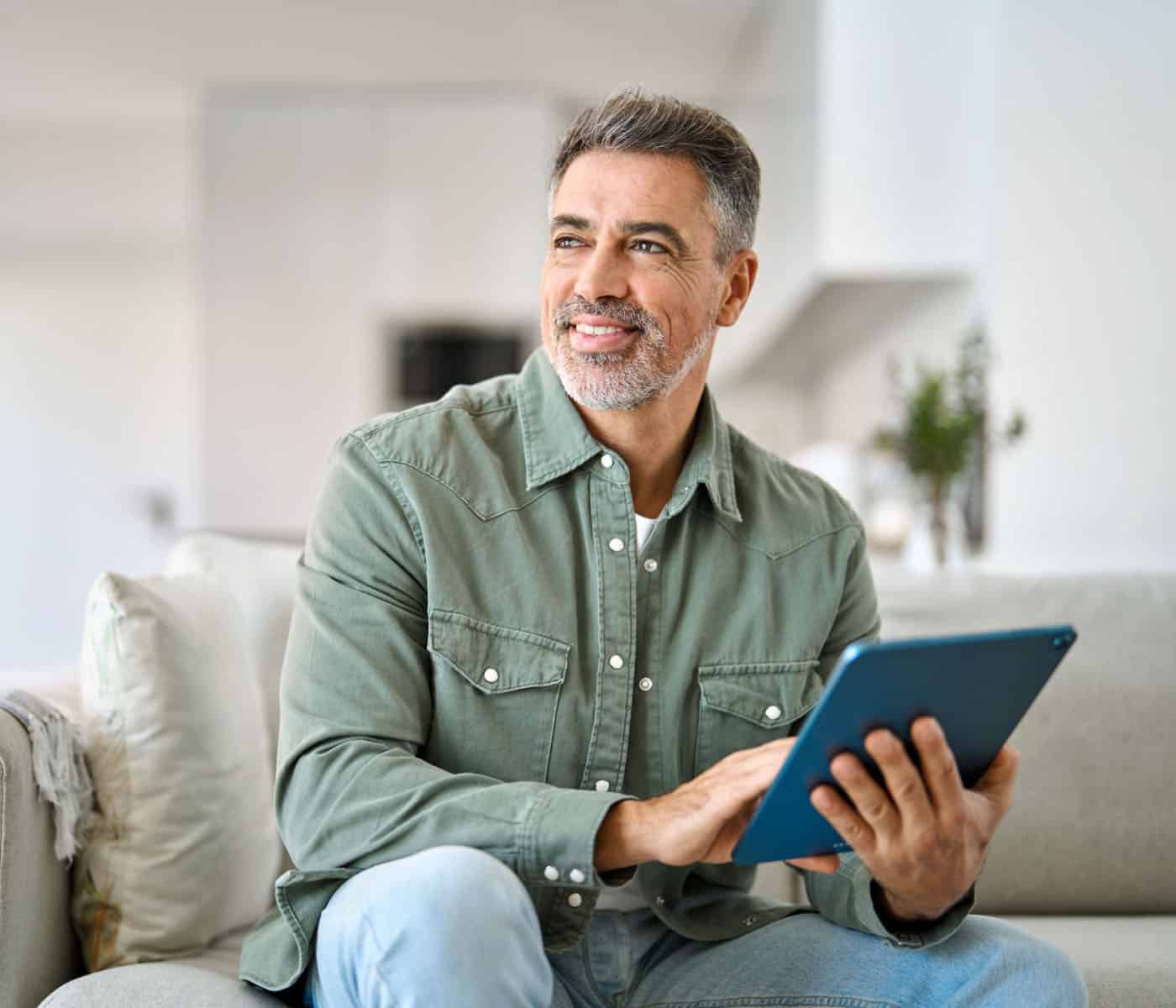 A man with salt-and-pepper hair and a beard sits on a couch, smiling while holding a tablet. Dressed in a green button-up shirt and jeans, he looks content in the bright, modern living room—perhaps browsing the latest tips for efficient heating.
