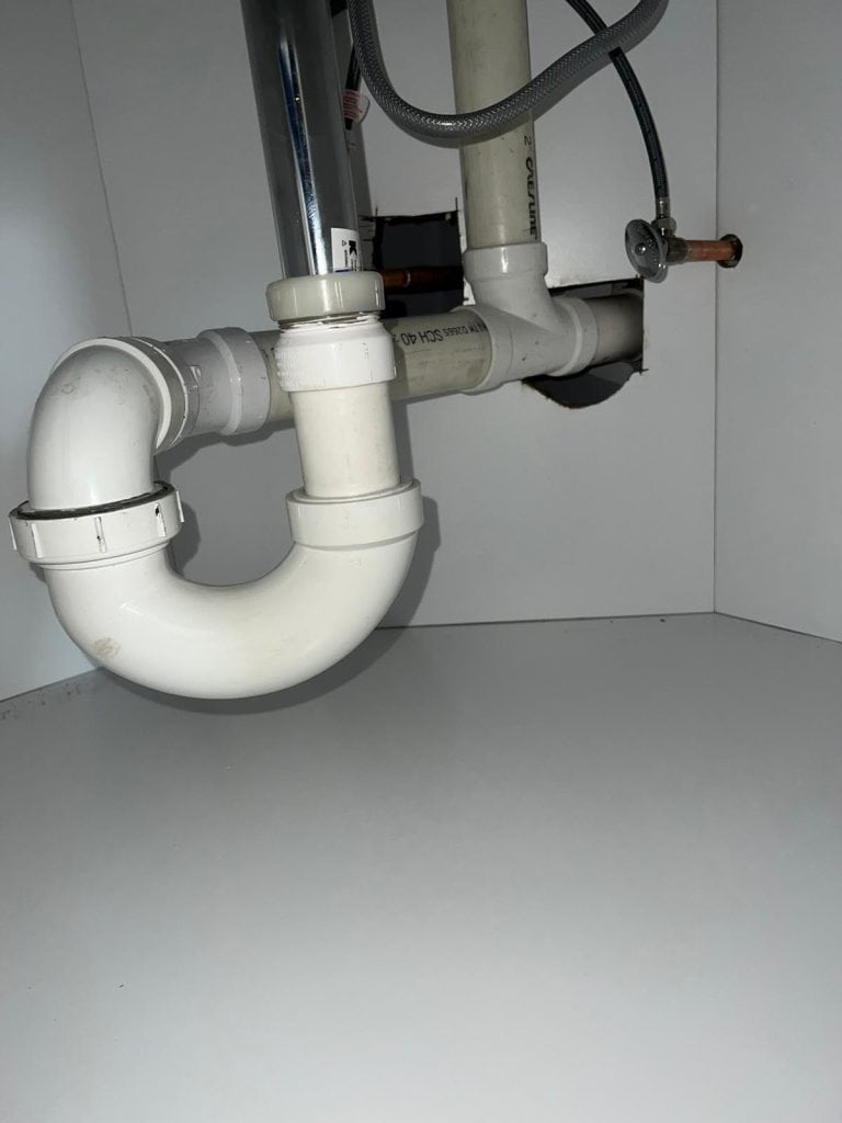 The image displays the plumbing setup under a sink, featuring white PVC pipes, joints, and a P-trap, seamlessly integrated with additional piping and a flexible metal hose. This efficient plumbing arrangement is housed within white cabinet walls.
