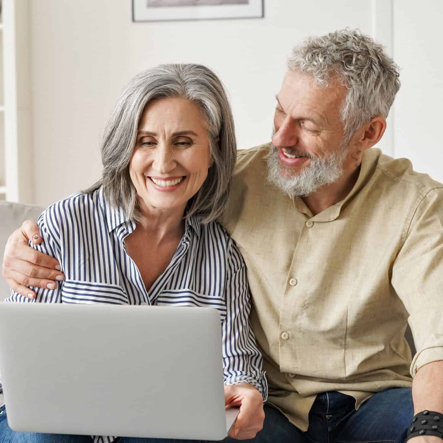 An older couple sits together on a couch, smiling and looking at a laptop. The woman wears a striped shirt, while the man in beige has his arm around her. They appear happy and relaxed in their bright living room, possibly searching for reliable plumbing services online.