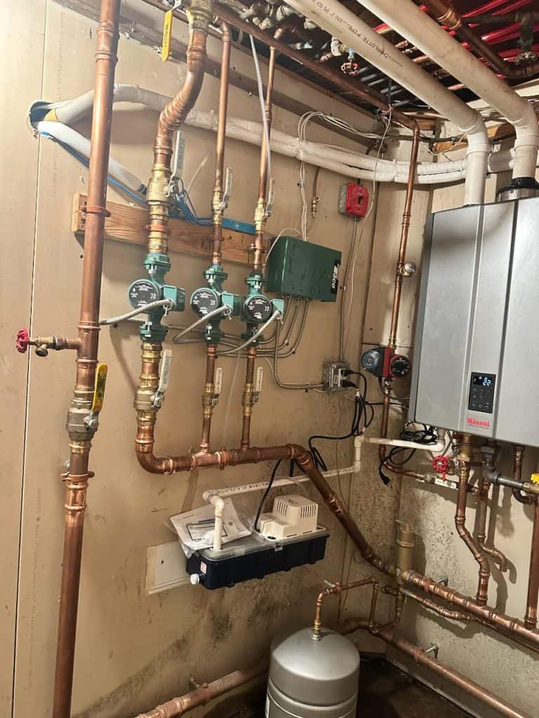The utility room features a water heater system and copper pipes, showcasing expert plumbing and heating work. Green valves complement the network of white and red pipes running along the ceiling and walls. A gray boiler is mounted on the right wall, with a small control box neatly attached nearby.