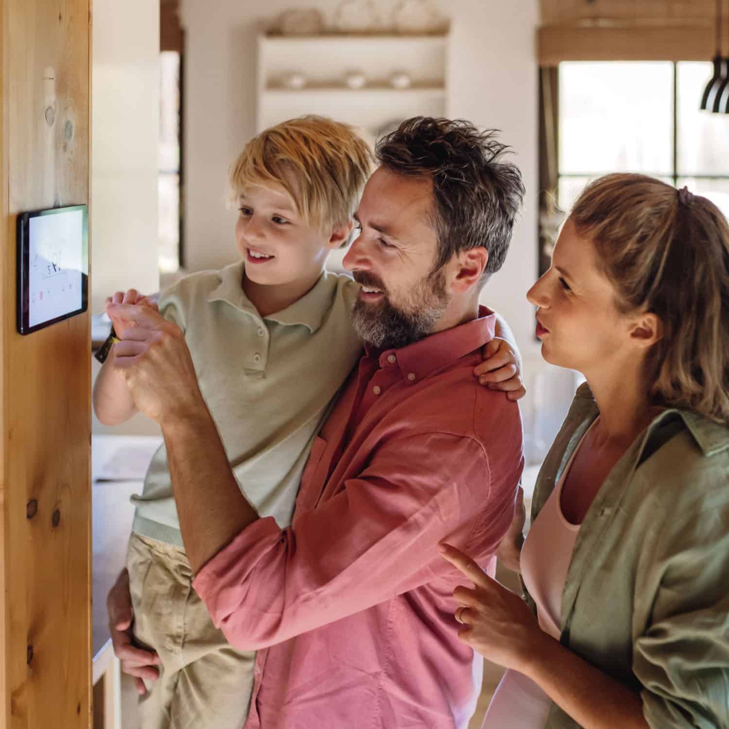A family of three, including a child and two adults, looks at a smart device mounted on a wooden wall in a cozy, well-lit room. They smile as they interact with the device, possibly adjusting their home's heating or checking on plumbing systems.