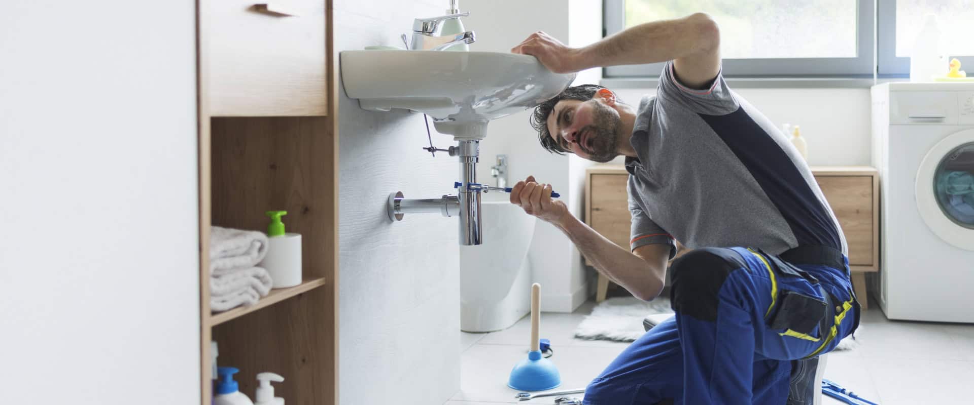A plumber in blue overalls and a gray shirt kneels in a bathroom, fixing a sink pipe with a wrench. Surrounded by plumbing tools, a plunger, and a washing machine, the scene highlights expert plumbing skills. Natural light streams through the window over a modern wooden cabinet.