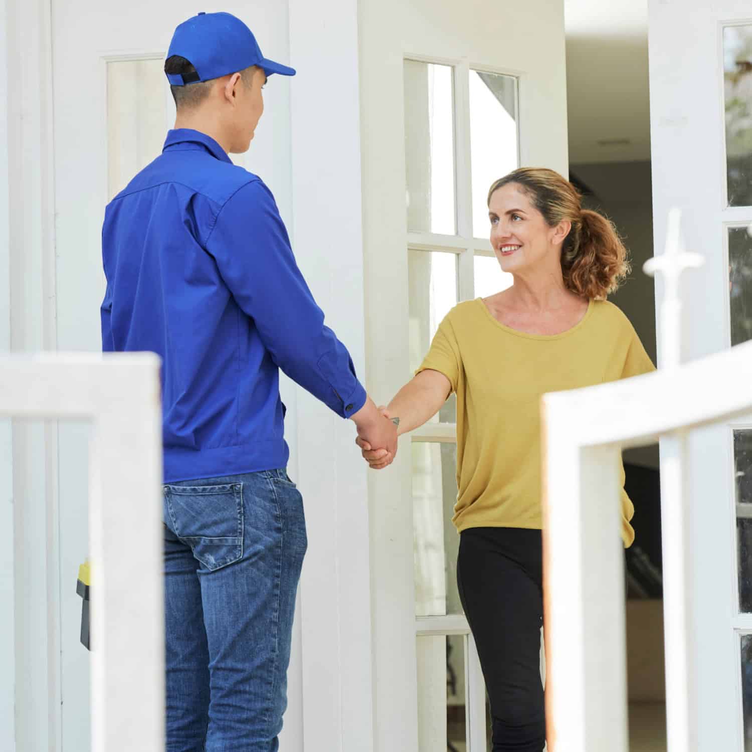 A person in a blue uniform and cap shakes hands with a smiling woman in a yellow shirt at her doorway, suggesting a plumbing & heating service call. The setting appears to be a residential entrance.