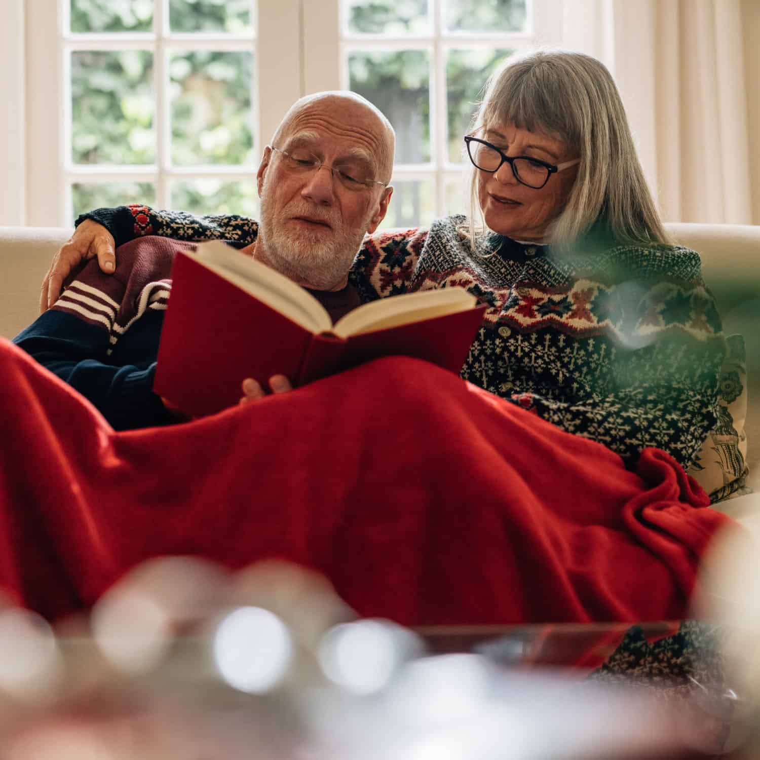 An elderly couple sits on a sofa, wrapped in a red blanket, reading a book together. Both wear cozy sweaters, while the gentle hum of the heating system ensures their comfort. The large windows reveal lush green foliage outside. The mood is warm and intimate.