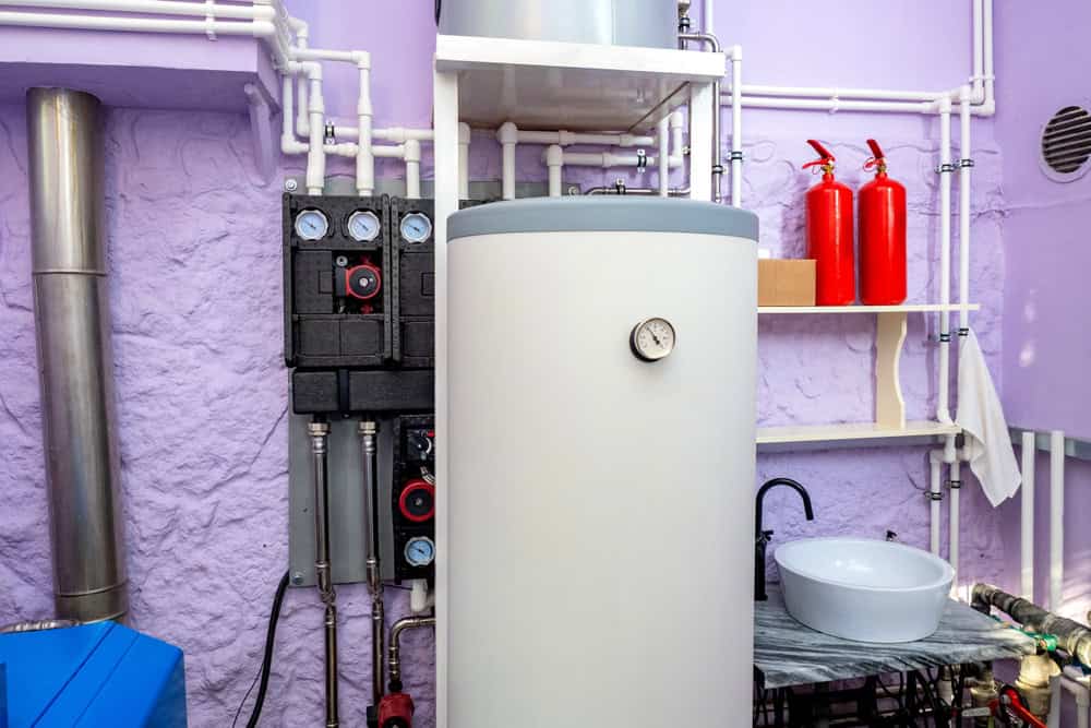 A utility room with a large white boiler, pressure gauge, pipes, and valves against a purple wall. Two red fire extinguishers are on a shelf above a white sink.