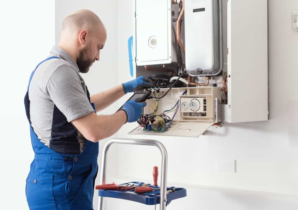 A technician in blue overalls is repairing a wall-mounted boiler. He is using tools from a nearby step stool and examining the wiring inside the open panel.