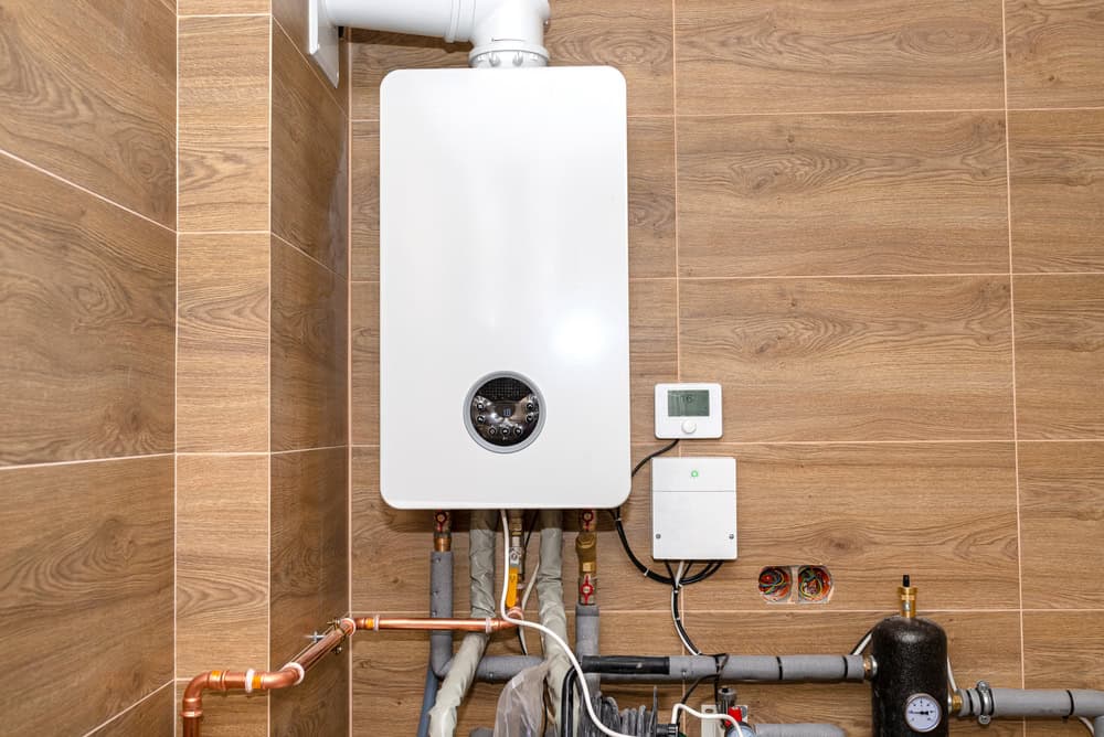 A modern white water heater is mounted on a wooden paneled wall. Below, various pipes and a black valve are connected. Nearby is a digital thermostat and a control box with a green light. The room has a neat and organized appearance.