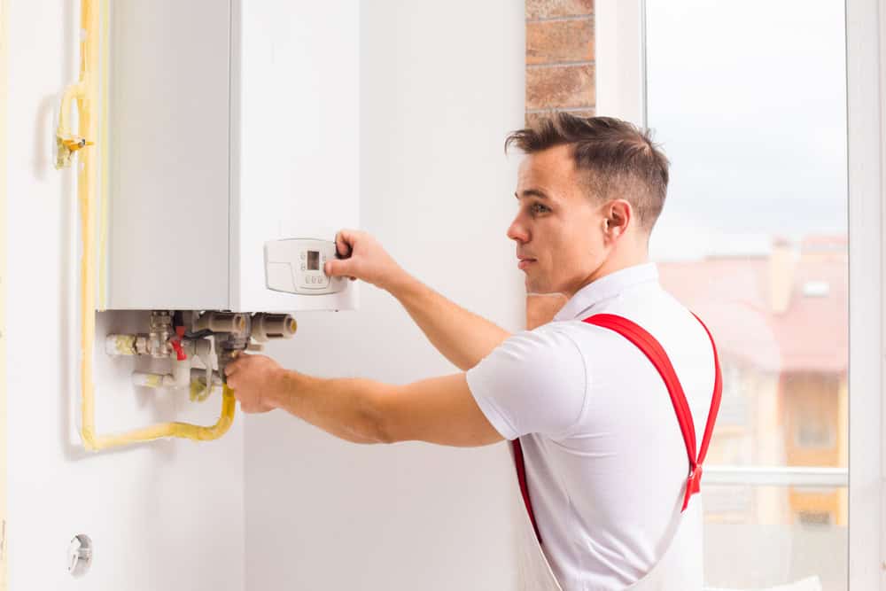 A technician in a white shirt and red suspenders adjusts a control panel on a wall-mounted device. He is standing by a window, with yellow pipes visible below the device.