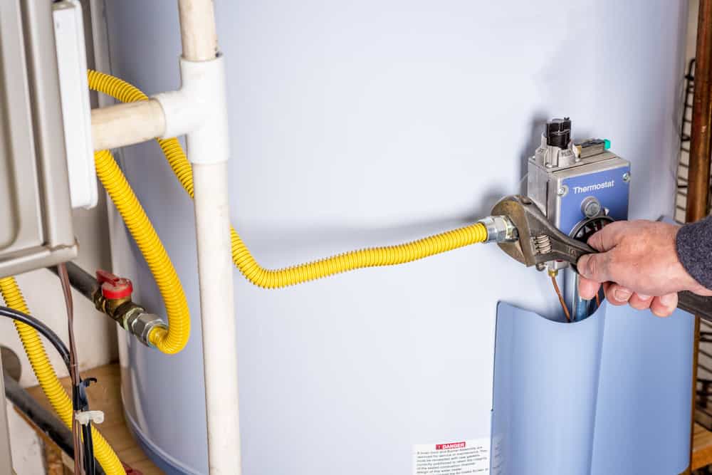 A person using a wrench to tighten a gas line connection on a water heater. The heater has a thermostat and yellow gas tubing. Pipes are visible in the background.