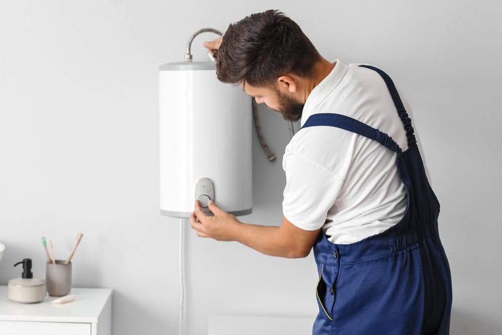 A man wearing blue overalls and a white t-shirt is adjusting a white wall-mounted water heater. He appears to be inspecting or repairing it in a home setting. The room is minimally decorated with a small table holding soaps and brushes.