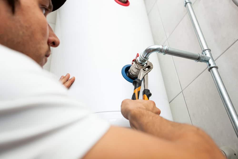 A person wearing a cap uses a wrench to adjust piping on a white water heater tank attached to a tiled wall. The image focuses on their hands and the plumbing work.