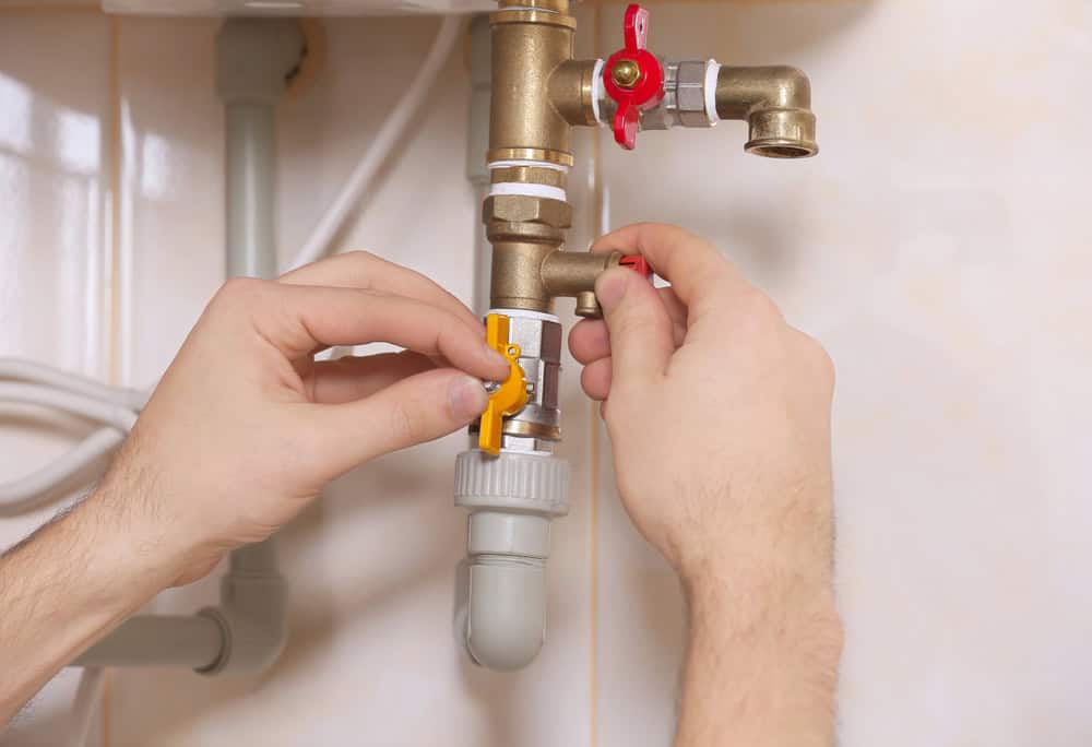Close-up of hands adjusting a yellow valve on a complex system of brass pipes with red levers in a plumbing setup. The background shows a tiled wall, suggesting an indoor setting.