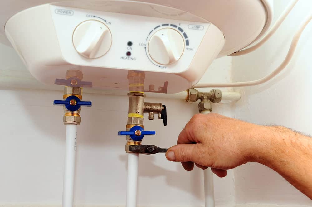 A person adjusts the plumbing beneath a water heater using a wrench. The water heater has two dials, labeled "Power" and "Temp," and several pipes with blue-handled valves connected to it.
