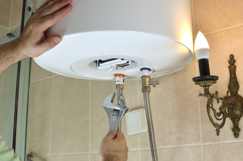 A person uses a wrench to adjust a pipe connected to a large white water heater mounted on a beige tiled wall. A vintage-style candle-shaped wall sconce is visible nearby. The hands focus on tightening or loosening the connections.