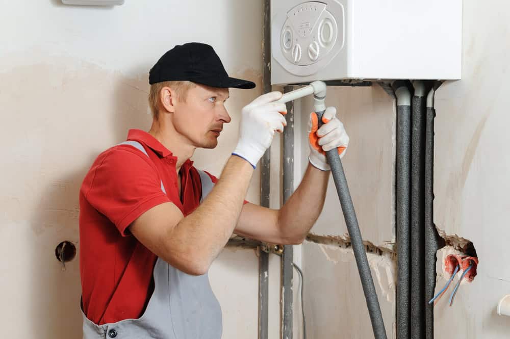 A worker in a red shirt, gray overalls, and black cap is installing or repairing a pipe connected to a white wall-mounted unit. He uses a wrench while standing in a partially finished room with visible wiring and pipes.