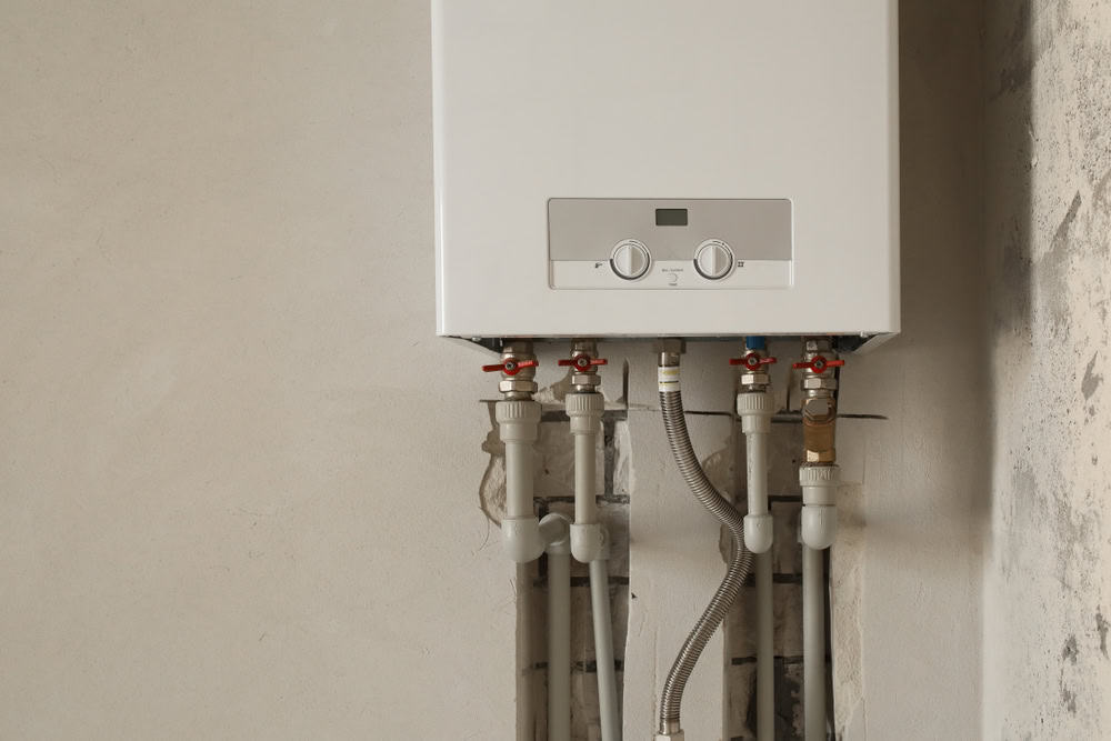 A wall-mounted white gas boiler with control knobs and a digital display. Multiple pipes are connected to the bottom of the boiler, seen against a rough, unfinished wall.