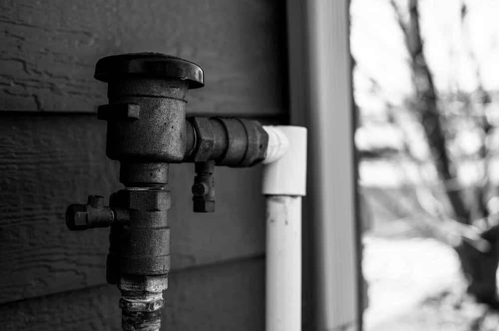 Black and white image of an outdoor water pipe with a dark valve, attached to a wall. The pipe is connected to a white vertical pipe. Blurred background with a hint of tree branches.