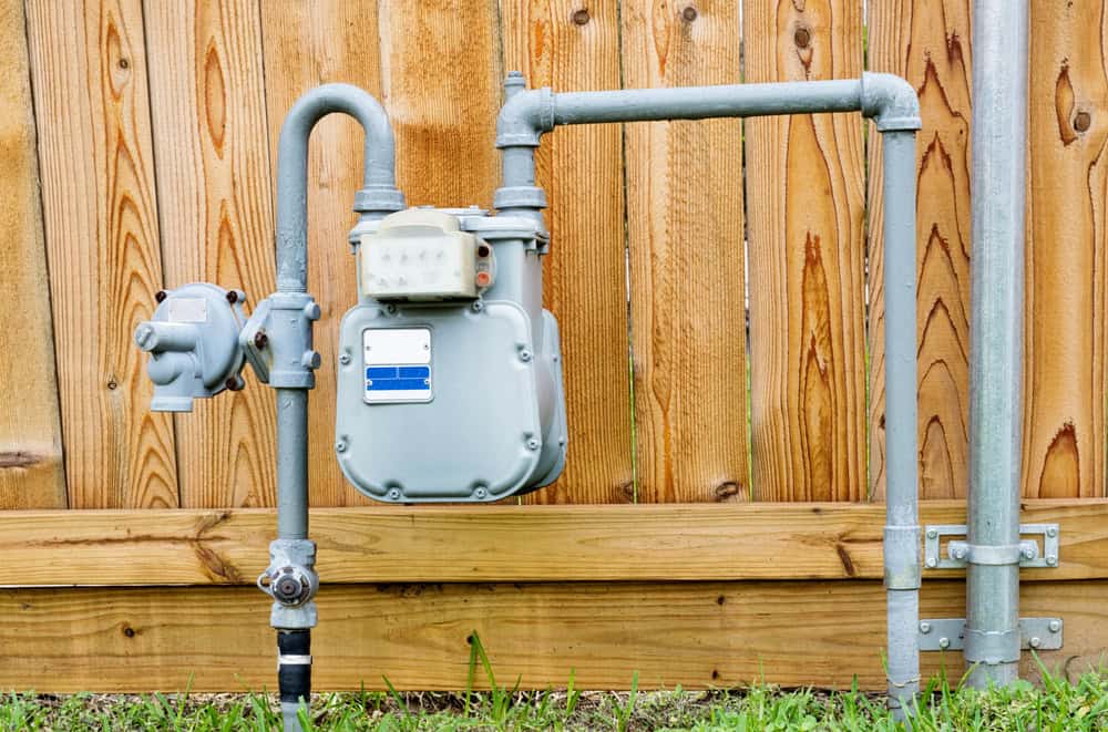 A gas meter with connected pipes is mounted on a wooden fence. The setup includes a vertical pipe leading to the meter and a horizontal pipe extending across the fence. The background shows a wooden fence and grassy area below.