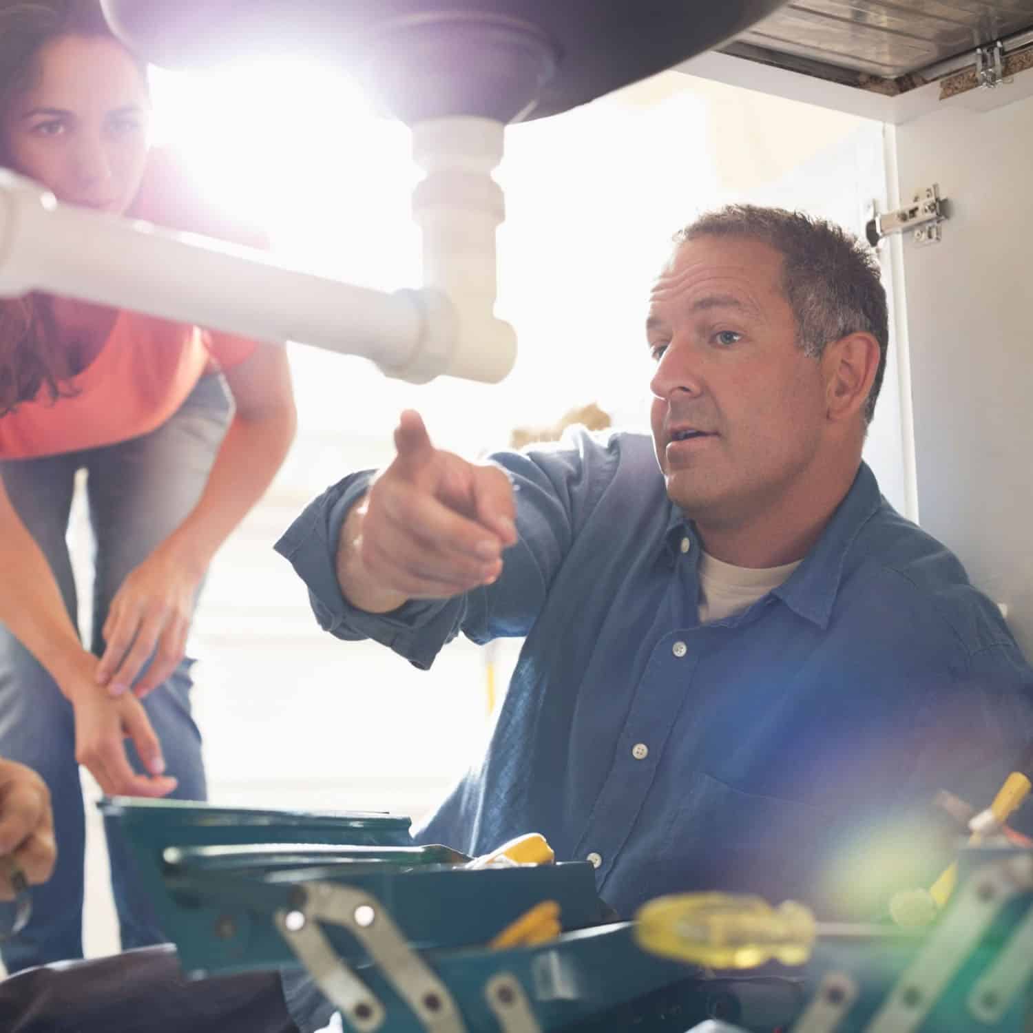 A man in a blue shirt works under a sink, pointing and explaining something to a woman in an orange shirt who is crouching nearby. A toolbox with various tools is visible in the foreground. Bright light shines in from behind them.