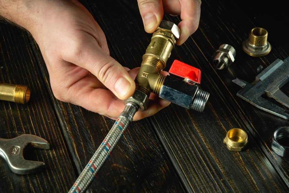 Hands assembling plumbing parts on a dark wooden surface, including a flexible hose, fittings, a wrench, and various connectors.