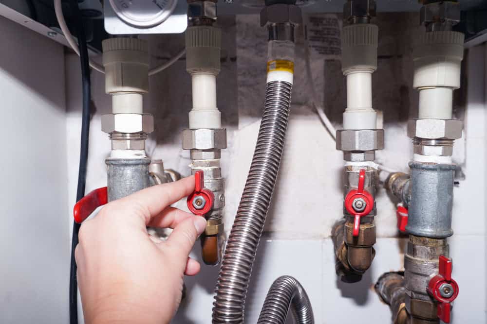 A hand is adjusting a red valve among a series of pipes and fittings, including a flexible metal hose, within a plumbing system. The setup is under bright lighting, with nuts and connectors visible.