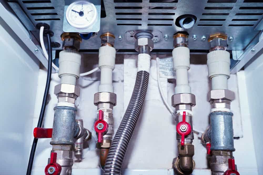 Close-up view of plumbing pipes and fixtures, featuring several metal pipes connected with valves and fittings. A gauge is visible at the top, indicating pressure measurements. The setup appears to be part of a heating or water system.