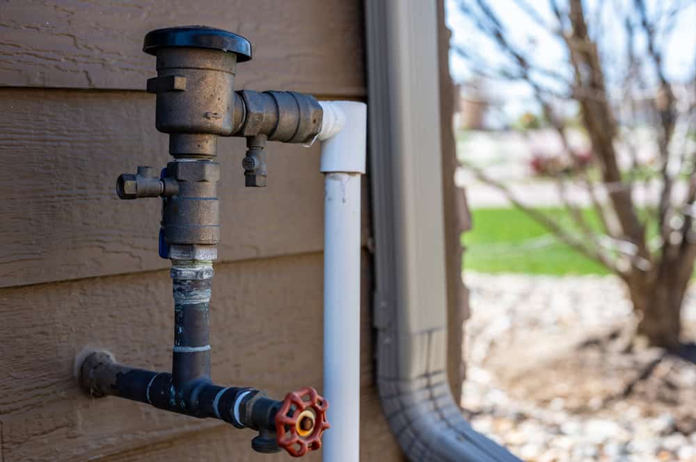 Close-up of an outdoor water spigot attached to a brown house wall. The spigot is connected to a series of pipes, including a vertical white pipe. There is a red shut-off valve and a black backflow preventer. Background includes blurred trees and grass.