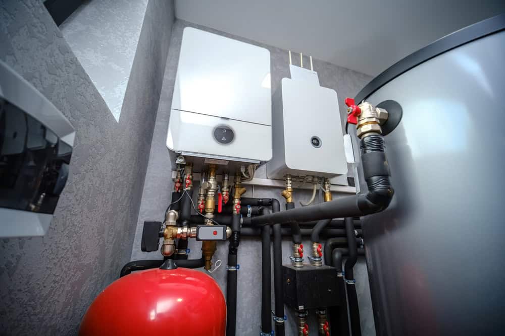 A modern boiler room setup with various pipes, valves, and two white heating units mounted on the wall. A red expansion tank is visible in the foreground, and a large cylindrical heating tank stands to the right.