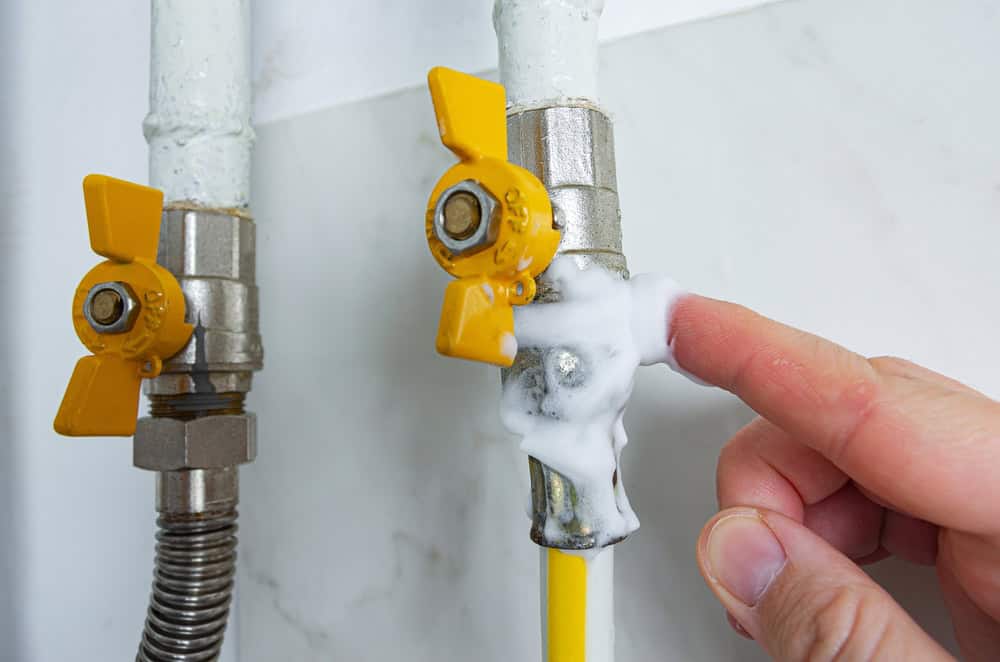 A hand applies soapy solution to a gas valve connection to check for leaks. The valve has a yellow handle, and the pipes are against a white tiled wall. Bubbles form at the connection point, indicating a potential leak.
