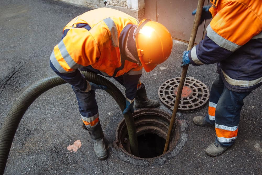 Two workers in orange safety uniforms and helmets are cleaning a street sewer. One is holding a hose while the other handles a pole, both positioned over an open manhole. The road surface around them is paved with asphalt.