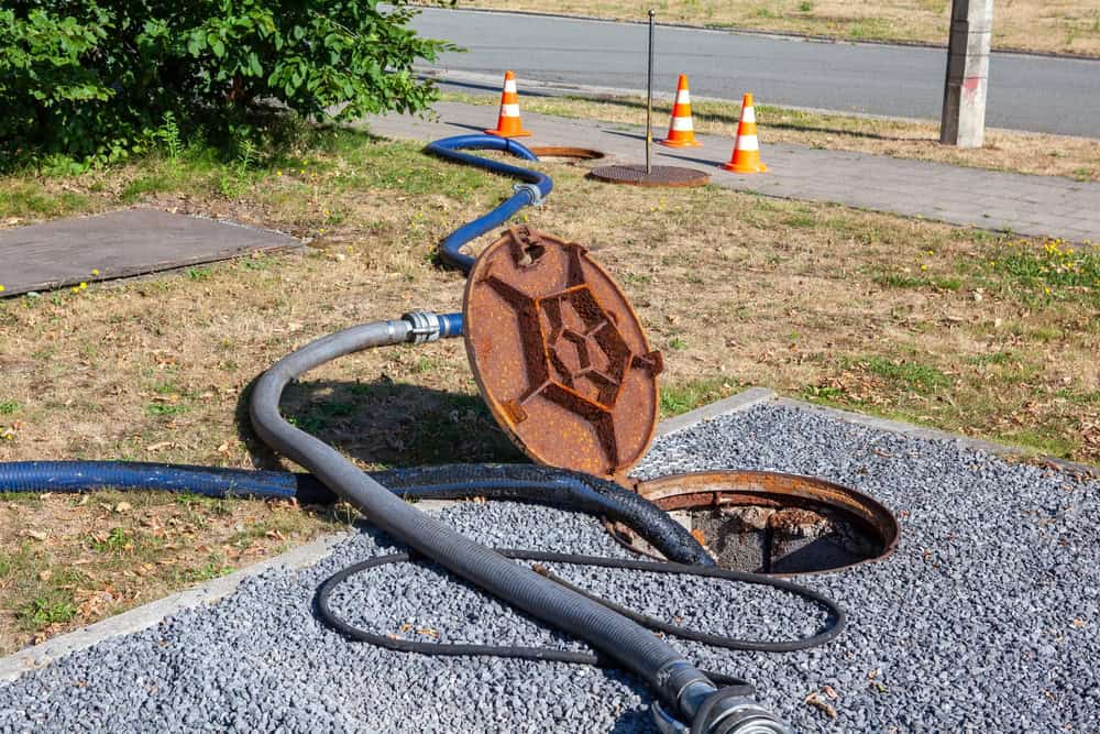 A sewer maintenance scene with an open manhole cover on the ground, connected to long hoses. Traffic cones are placed behind the opening for safety. A patch of grass and a road are visible in the background.