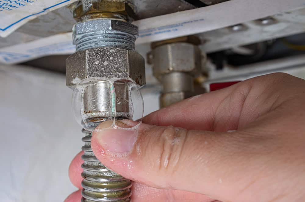 A hand checks for a gas leak by applying soapy water to a metal pipe connection. Bubbles are visible, indicating a potential leak. The setting appears to be underneath a sink or appliance.