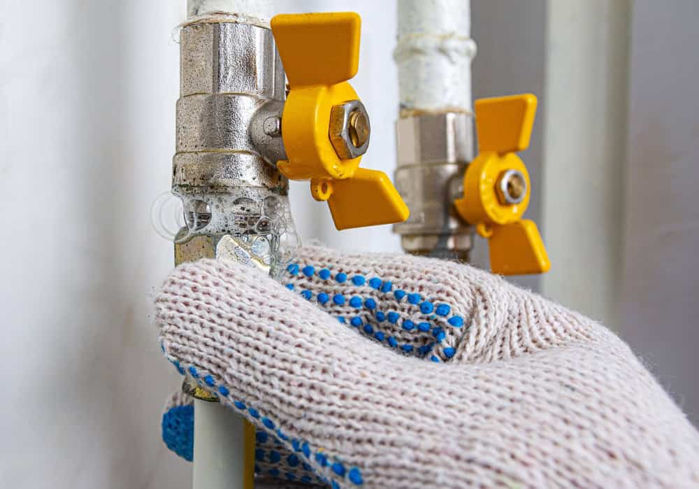A gloved hand is adjusting a yellow valve on a metallic pipe. The glove is white with blue dots. Two valves are visible, both with yellow handles, on a white and silver pipe system.