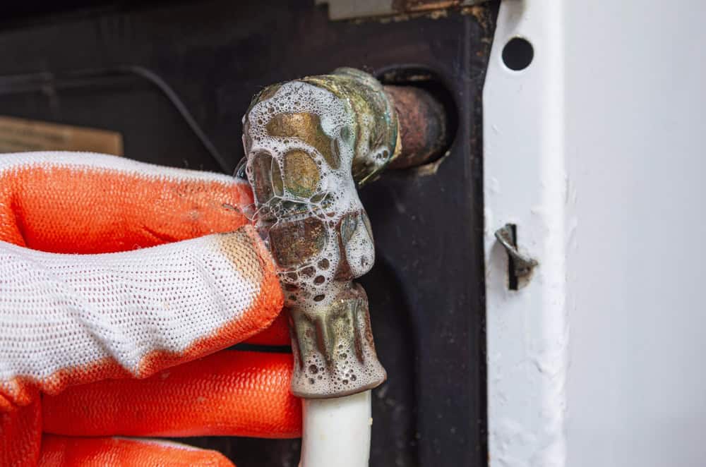 A person wearing an orange glove checks a pipe connection for leaks using soapy water. Bubbles form on the joint, indicating a possible gas or air leak. The setting appears to be an outdoor maintenance area.