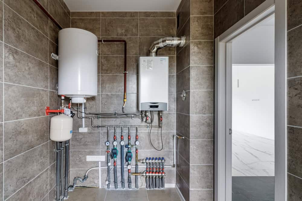 A utility room with gray tiled walls, featuring a white water heater tank, pipes, and meters mounted on the wall. An open doorway leads to a bright, adjacent room with white walls and a marble-like floor.