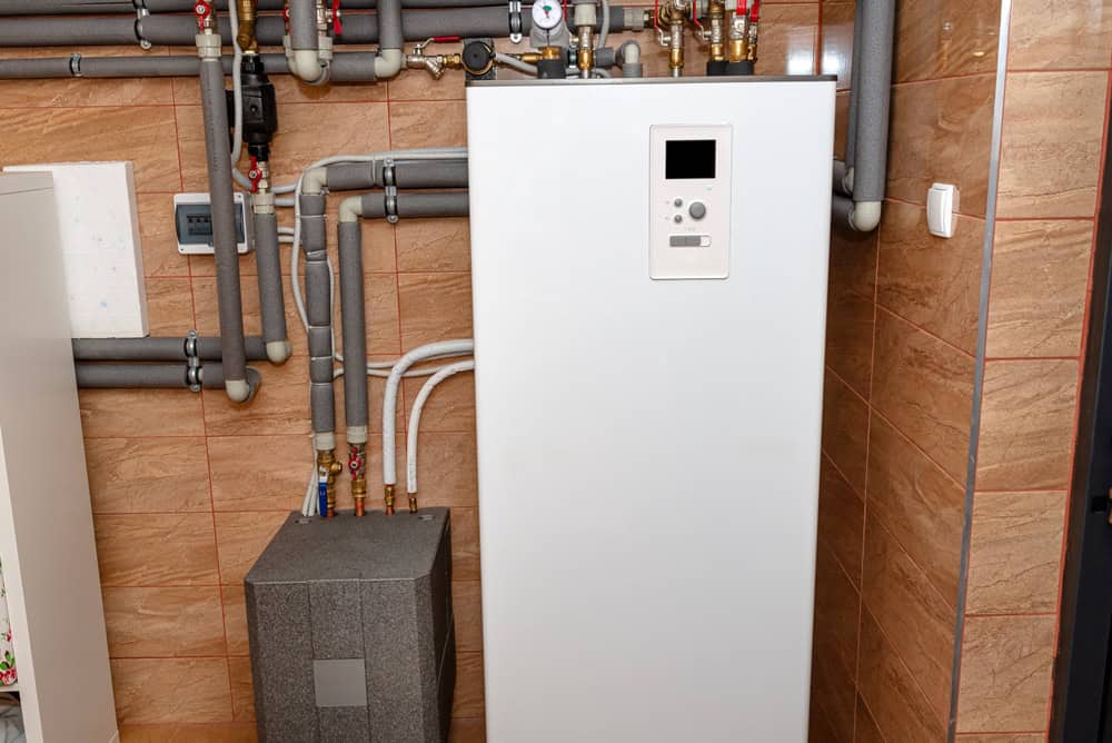 A modern white boiler unit is installed against a tiled wall, connected to various pipes and valves. The setup is in a utility room, with copper and insulated pipes visible, indicating part of a heating system.