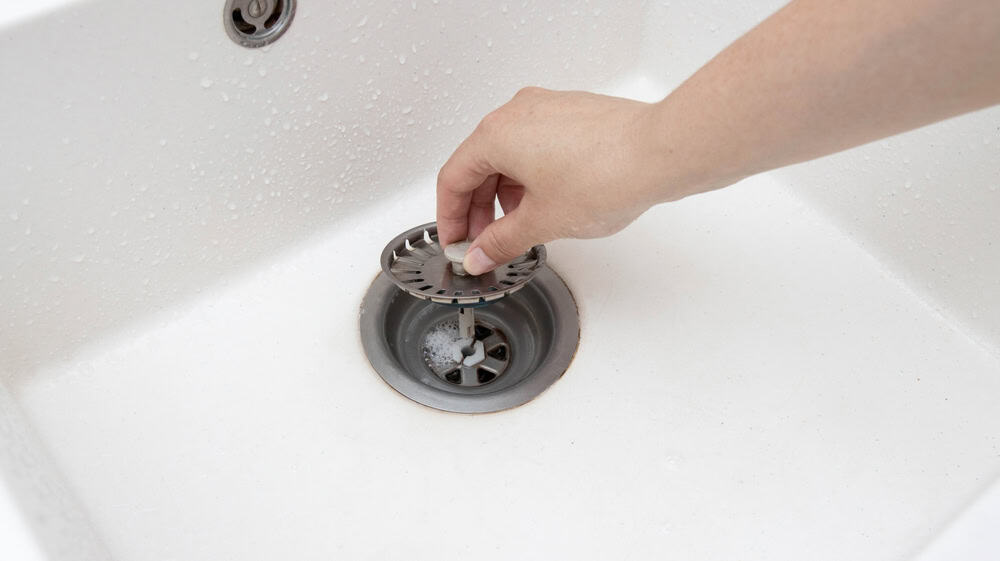 A hand lifts a metal sink strainer from a white kitchen sink. Water droplets are visible around the sink, suggesting cleaning activity.