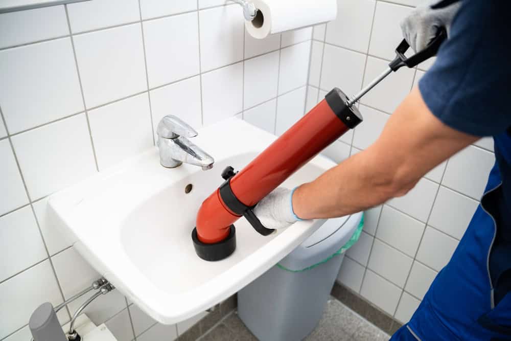 A person in blue overalls and white gloves is using a red plunger to unclog a white sink in a tiled bathroom. A roll of toilet paper is visible in the background.