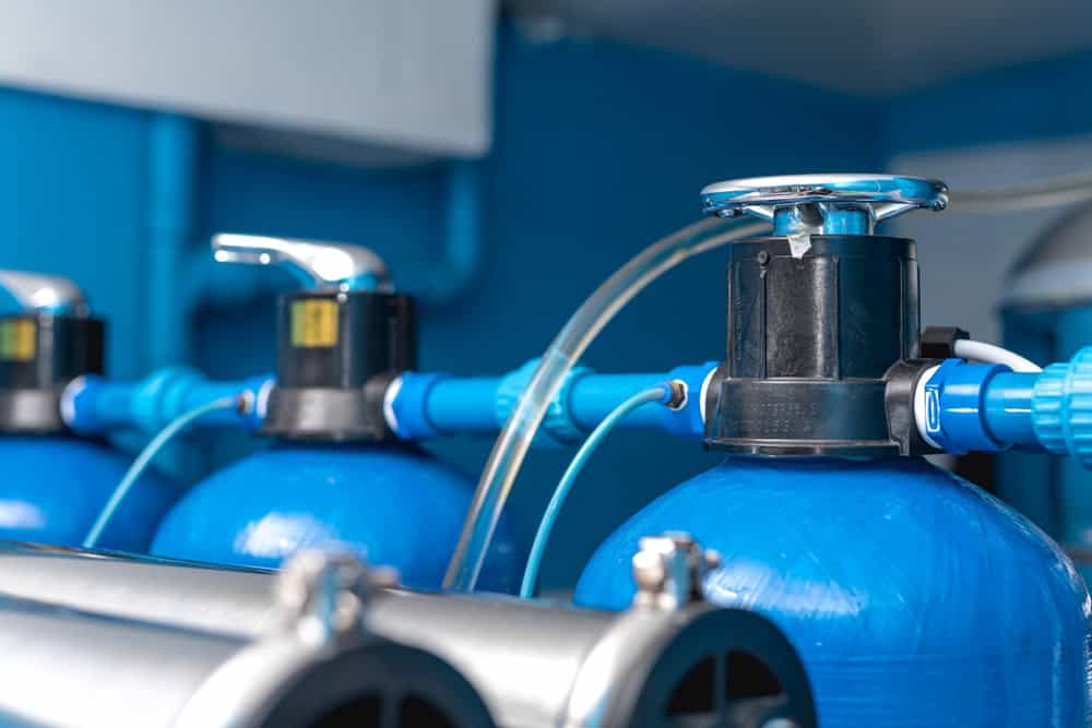 Close-up of industrial water purifiers featuring blue tanks, pipes, and valves. The equipment is arranged in a row, with metallic components and tubes connected for water filtration. The background is a blurred blue shade.