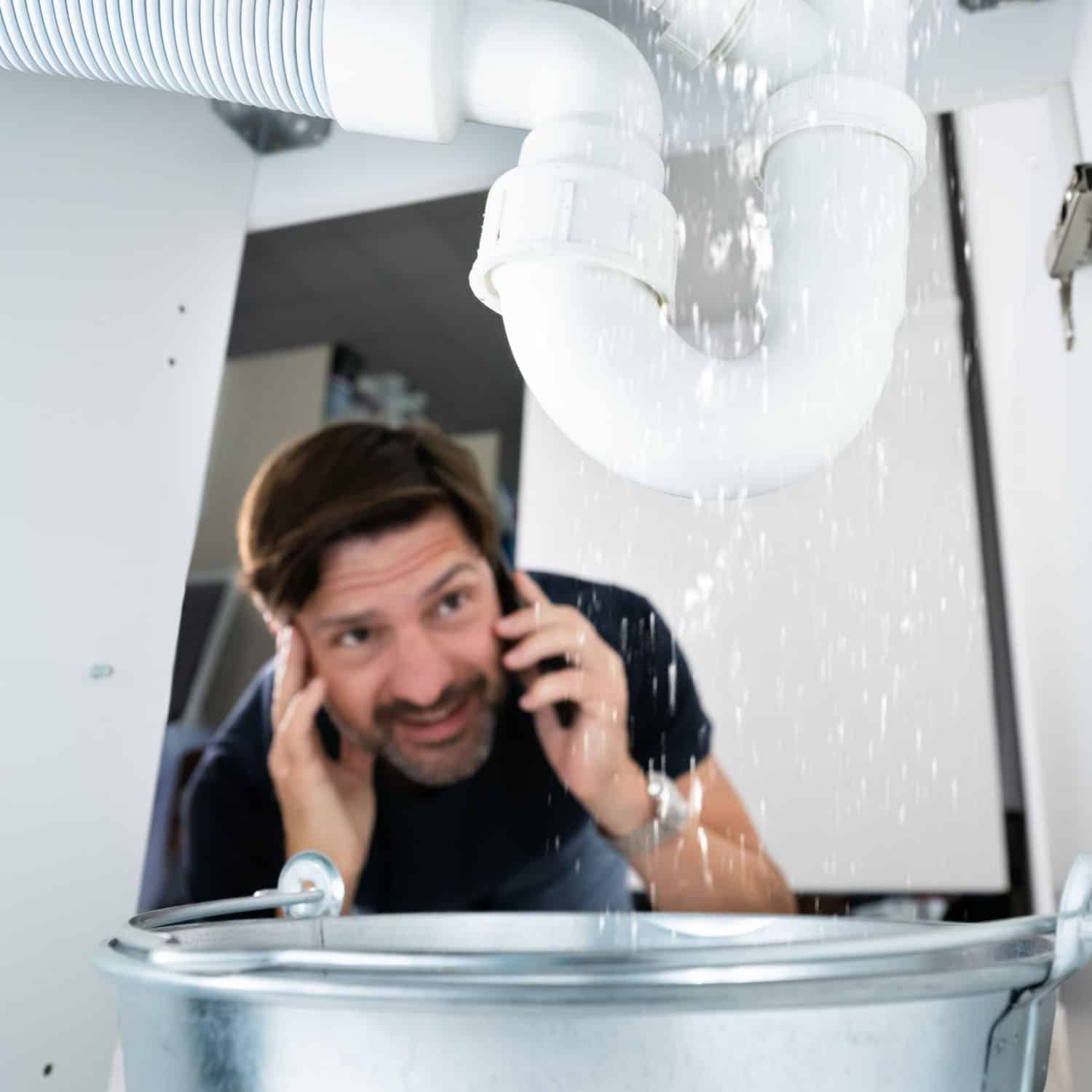 A man is crouching under a sink, looking up at a leaking pipe while talking on the phone. Water is spilling into a bucket placed below the pipe. The setting appears to be a kitchen or bathroom.
