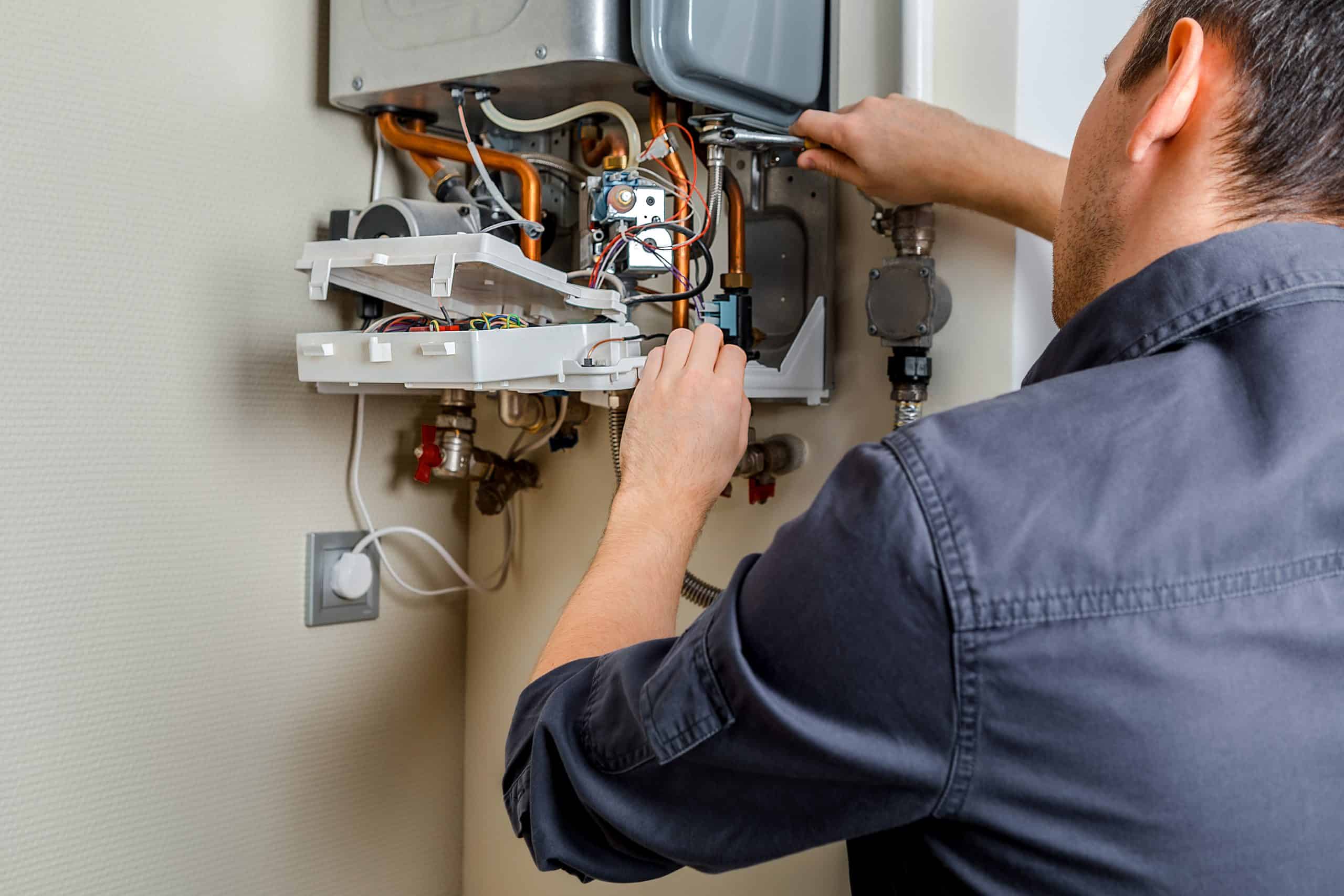 A person in a dark shirt, likely from a heating company in Nassau County NY, repairs a wall-mounted boiler with open panels and exposed wiring. They diligently adjust components inside the unit with tools, suggesting some maintenance or troubleshooting in a home setting.