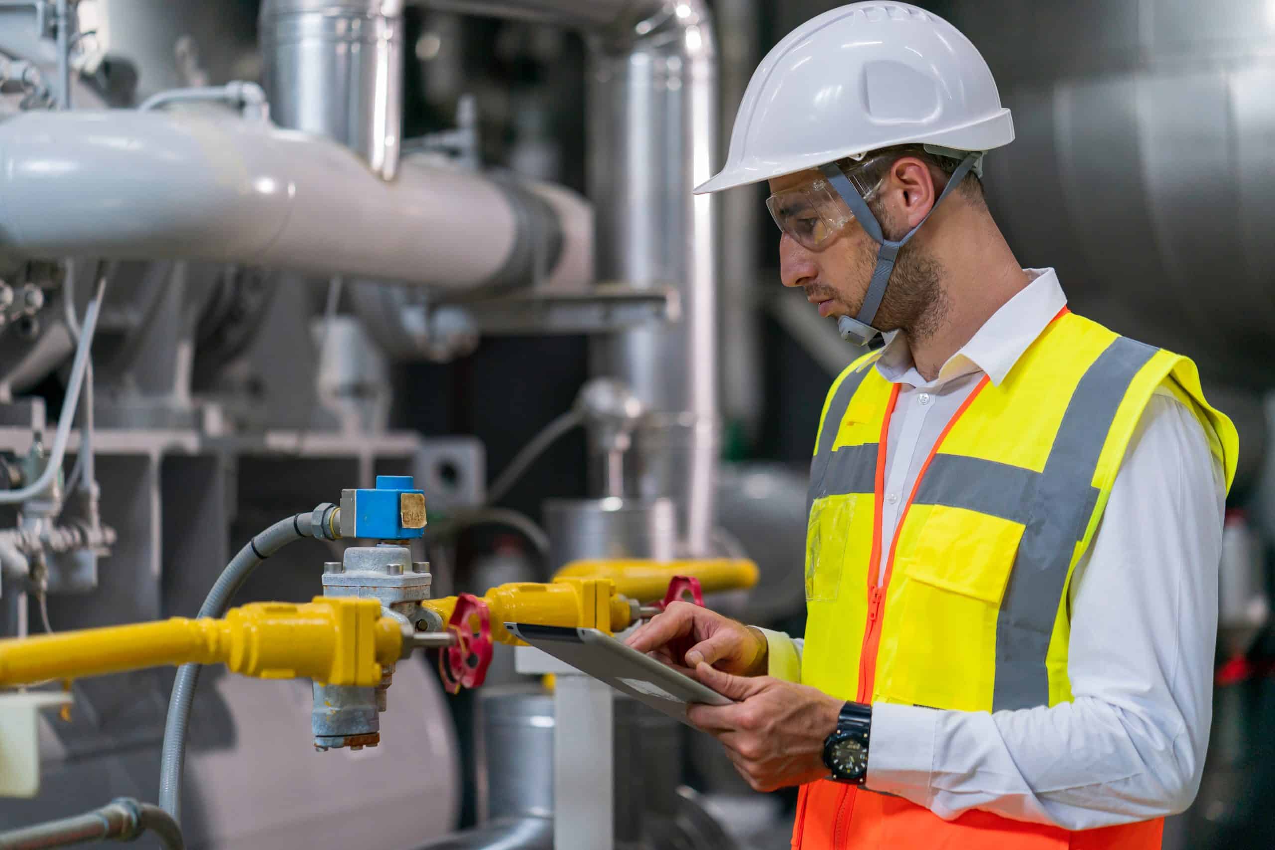 A man in a white hard hat and yellow safety vest is inspecting industrial equipment for a heating company in Nassau County, NY. Holding a tablet, he stands near machinery with pipes and valves in the factory setting, ensuring everything runs smoothly.