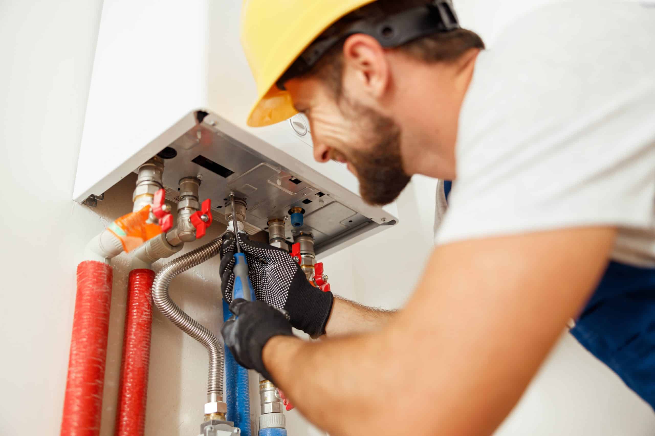 A plumber wearing a yellow hard hat and gloves is using a wrench to adjust pipes connected to a wall-mounted water heater. The pipes are red and blue, indicating hot and cold water lines. The plumber appears focused on his task.