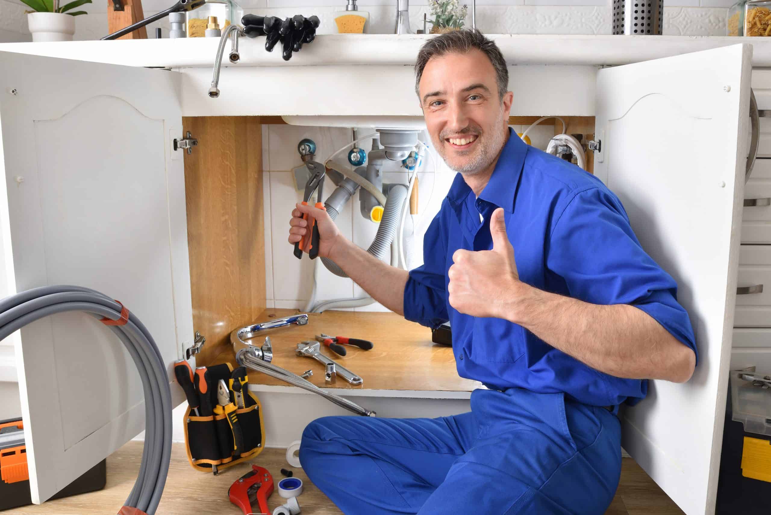 A plumber in a blue uniform kneels before an open sink cabinet, holding a screwdriver and giving a thumbs-up. Surrounded by scattered tools, he looks pleased with his work, embodying the reliable residential plumbing repairs Nassau County NY residents trust.