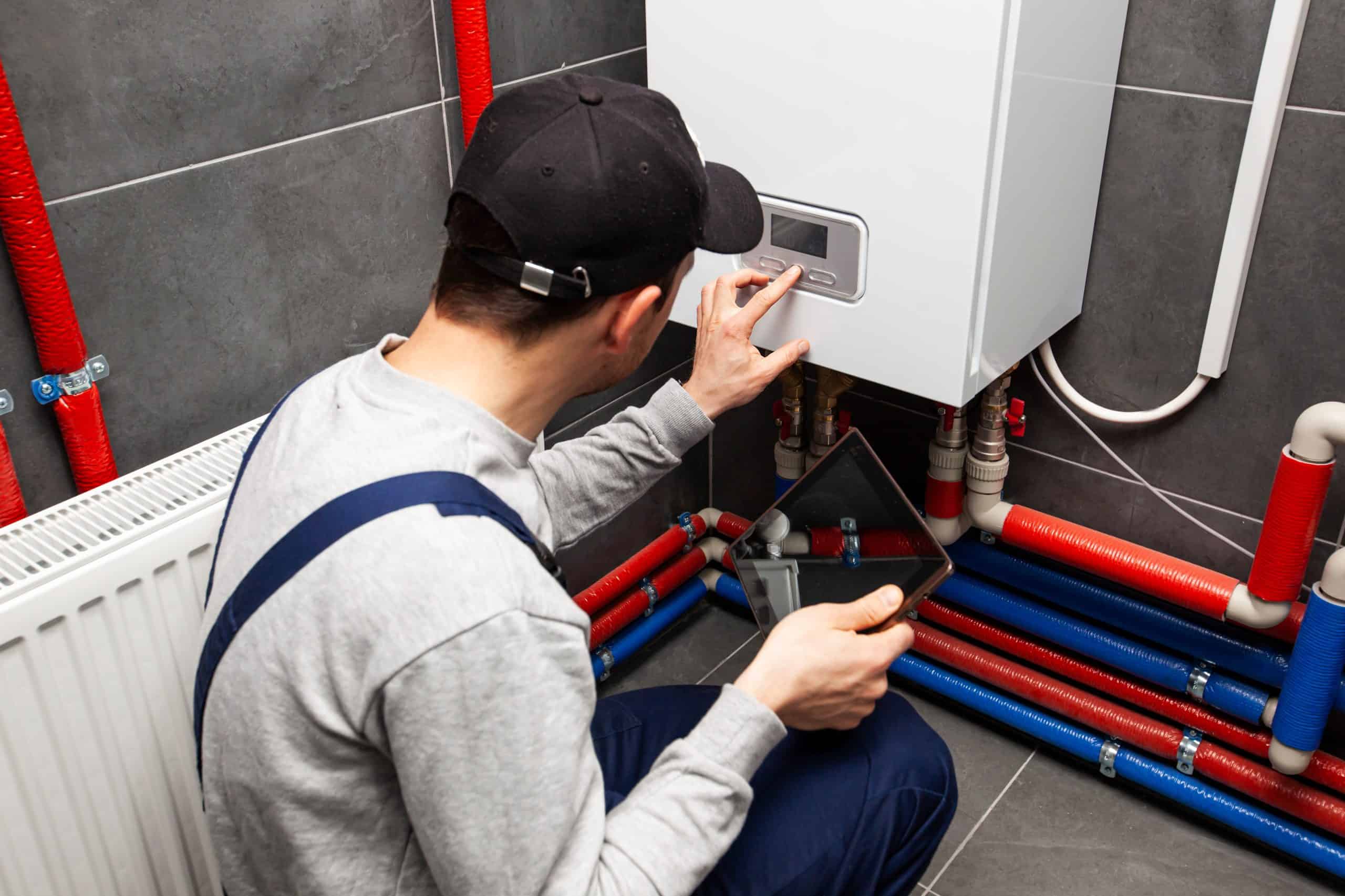 A person wearing a cap and overalls is kneeling in front of a white boiler, adjusting its controls with one hand while holding a tablet in the other. Red and blue pipes are visible, and a radiator is on the left.