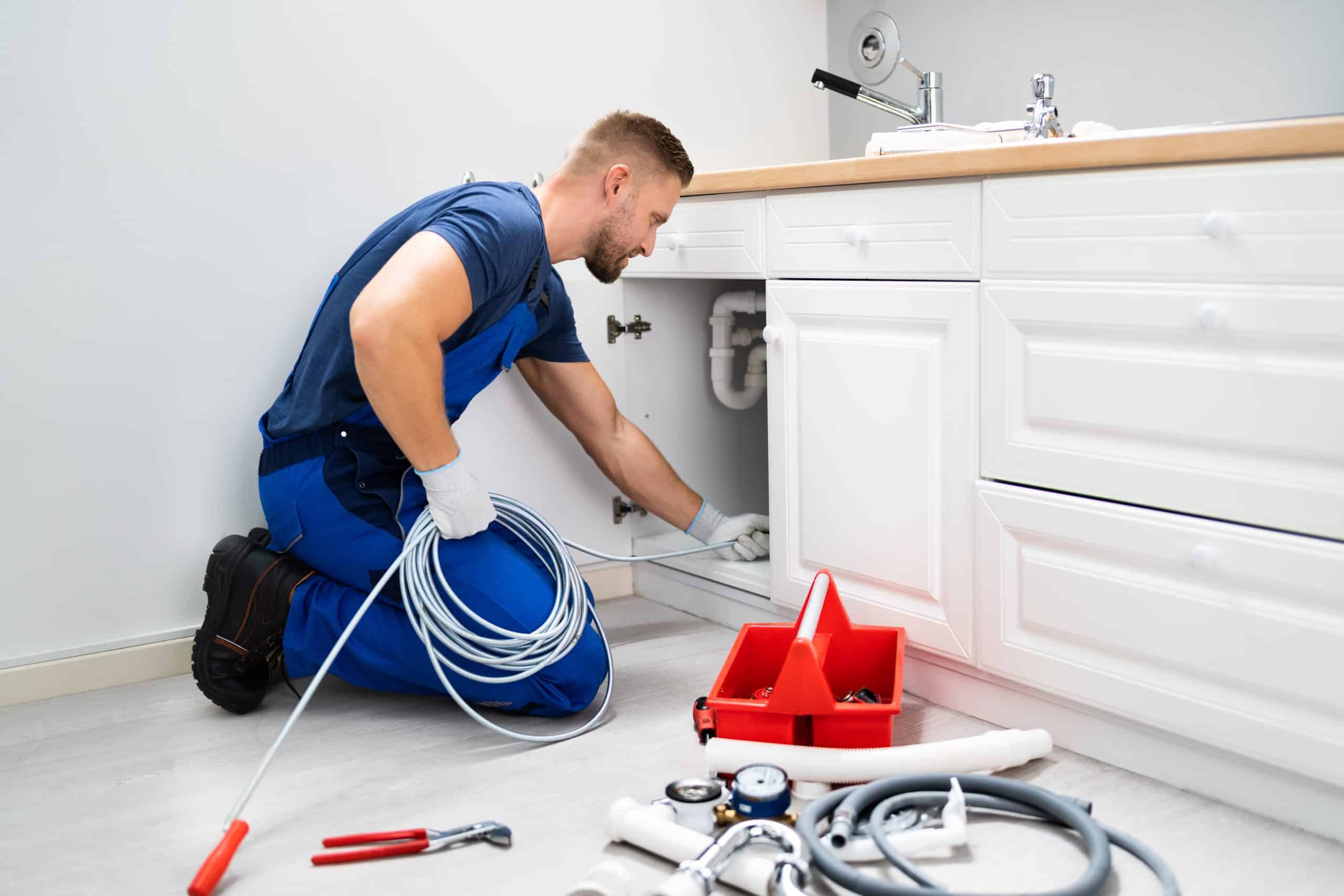A plumber in blue overalls is kneeling on the floor, examining pipes under a white kitchen sink. He's holding a coil of cable. Nearby, a red toolbox brims with tools—expertly handling residential plumbing repairs in Nassau County, NY, to ensure everything flows smoothly.