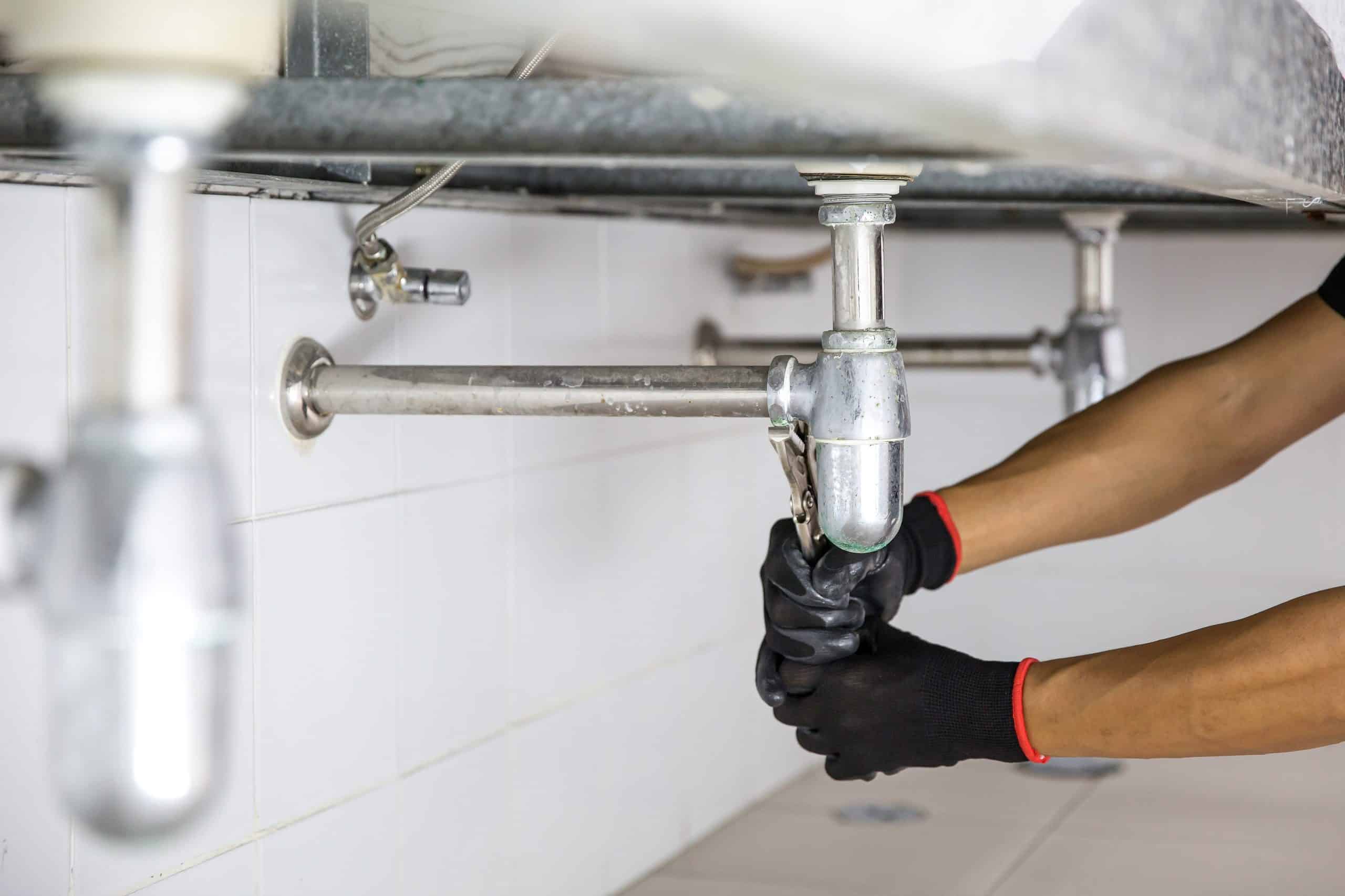 A person wearing black gloves is repairing a pipe under a sink. They are using a wrench to tighten or loosen a pipe fitting, in a setting with white tiled walls.