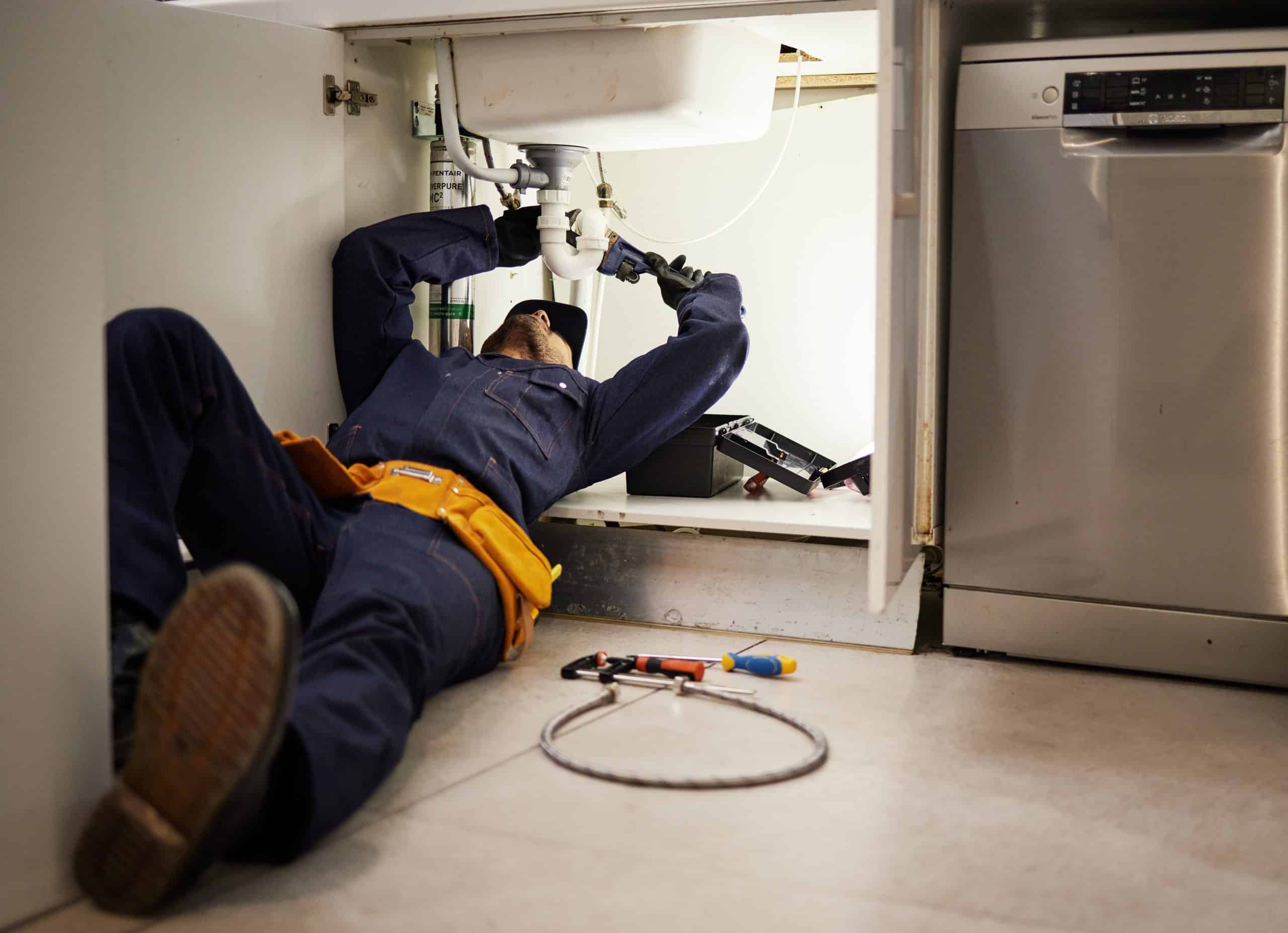 A plumber in navy overalls lies on his back under a kitchen sink, engaged in repair work. Various tools are scattered on the floor, including a wrench and pliers. A dishwasher is visible to the right.