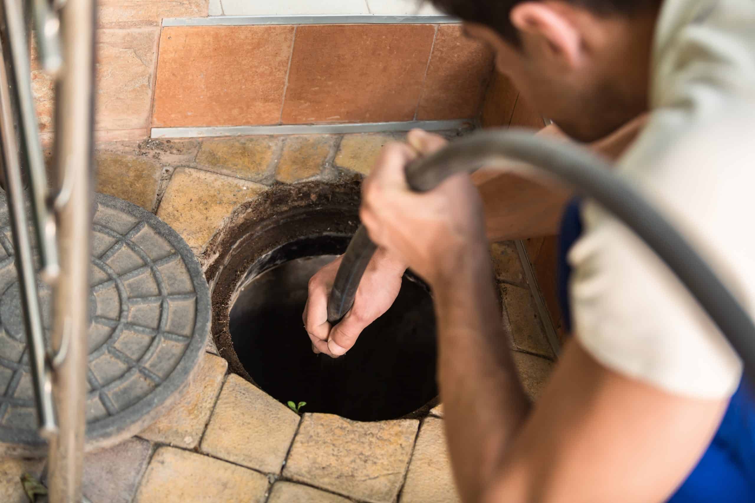 A person is using a hose to clean or inspect a drain on a tiled floor. The manhole cover is partially visible beside the open drain. The person is wearing a blue outfit.