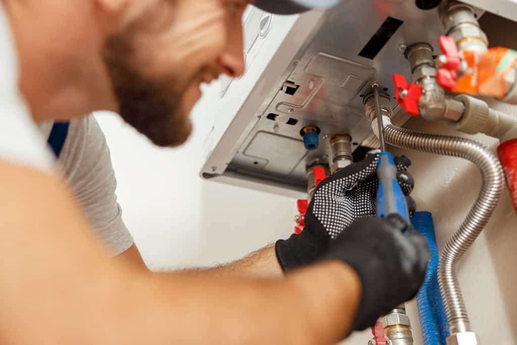 A person is focused on repairing or installing a piece of machinery or equipment. They are tightening a bolt using a wrench, wearing safety gloves, and surrounded by various pipes and fittings. The image suggests a plumbing or maintenance task.